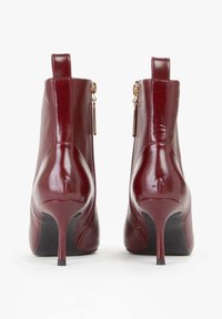 Stivaletti in pelle patent bordeaux con texture liscia, punta affusolata e tacco a spillo sottile. Presentano un dettaglio con zip in tonalità oro.