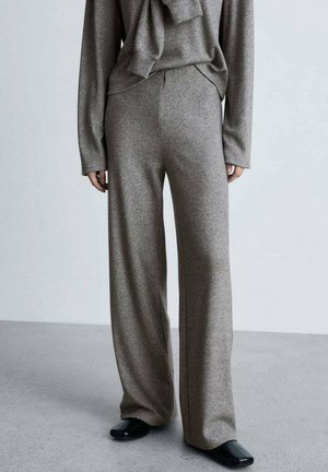 Person iført løstsiddende grå strikkede bukser, kombineret med matchende langærmet top og sorte sko med firkantet tå, stående på gråt gulv.