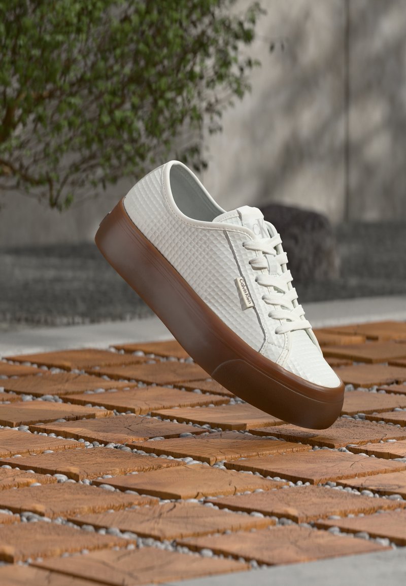 Baskets blanches texturées Calvin Klein avec semelle marron flottant au-dessus d'un carrelage en bois et d'un sol en galets en plein air.