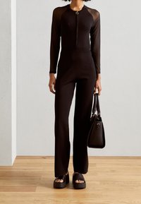 Combinaison noire à manches longues avec une fermeture éclair, accents transparents aux épaules, associée à des sandales à plateforme épaisses noires et un sac à main noir.