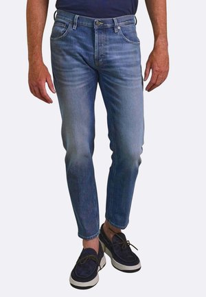 Uomo che indossa jeans slim fit blu e scarpe casual in suede blu scuro con suole bianche, in piedi davanti a uno sfondo chiaro e semplice.