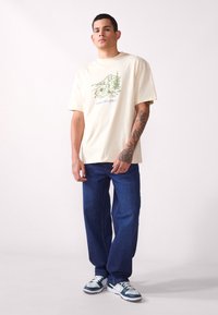 Camiseta de algodón crema con gráfico de montaña verde y texto "PHOENIX 112.0740", combinada con jeans de mezclilla azules y zapatillas multicolores.