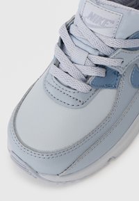 Svetlo modra Nike tenisica z gladkim usnjem, okroglo prstno območje, strukturirani nadstandardski deli in ravne vezalke. Ima izstopajoč logotip na ježku.