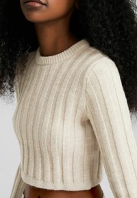 Beige ribbad stickad tröja med rundad hals och kortad design. Tyget ser mjukt ut med en texturerad yta som framhäver vertikala ränder.