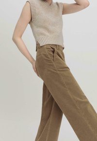 Beige pletený top bez rukávů s véčkovým výstřihem a žebrovaným lemem, kombinovaný s hnědými kordovými kalhotami s vysokým pasem a širokými nohavicemi, které mají přední kapsy.