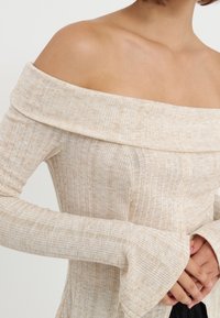 Beige off-shoulder topp med et teksturert strikkemønster, lange ermer og utstående mansjetter; glatt stoff med subtile variasjoner i farge.