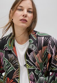 Crna jakna na zatvaranje s rajfešlusom s živopisnim uzorcima lišća u ružičastoj, zelenoj i narančastoj boji, složena preko bijelog majice s kratkim rukavima, s visokim ovratnikom.