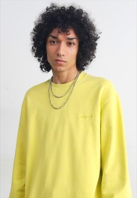 Gul sweatshirt lavet af blødt stof, med runde halsudskæring, lange ærmer og et diskret logo på brystet. Inkluderer lagdelte sølvkæder.