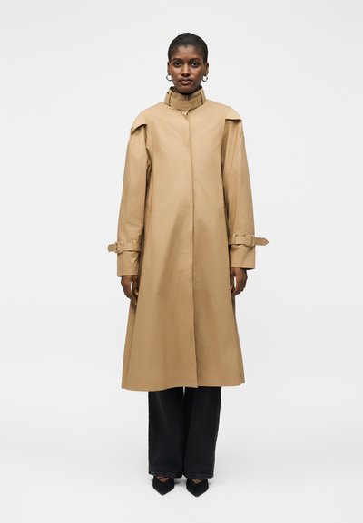 Femme portant un long trench-coat beige à col montant, avec des brides à boucle sur les manches, un pantalon noir à jambes larges et des chaussures noires pointues, debout sur un fond blanc.