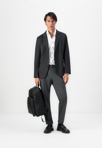 Mann, der gegen eine weiße Wand steht, in einem karierten Blazer, weißem Hemd, grauen Hosen, schwarzen Schuhen und der einen schwarzen Rucksack am Griff hält.