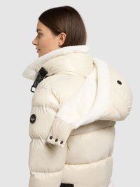 Kurtka puchowa w kremowym kolorze z kołnierzem z imitacji baranka. Posiada mankiety na guziki, boczne zamki błyskawiczne oraz błyszczące wykończenie. Logo na rękawie jako akcent.