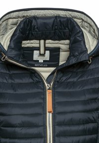 Marineblau gesteppte Jacke mit beiger Fütterung, sichtbarer Reißverschluss mit braunem Zipper, und "Camel Active Recycled"-Etikett im Kragen.
