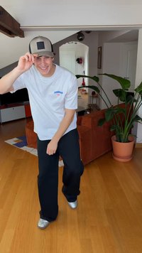 Camiseta blanca con logo azul de Adidas, pantalones negros de corte relajado y zapatos plateados. De fondo, un sofá marrón y una planta verde.