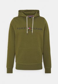 Olive green hoodie with front pocket, white drawstrings, and "Tommy Hilfiger Est. 1985" logo across the chest with a small flag emblem.