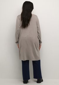 Tunica lunga grigia con maniche lunghe e vestibilità comoda, realizzata in materiale morbido. Abbbinata a jeans blu a gamba larga e stivali neri.