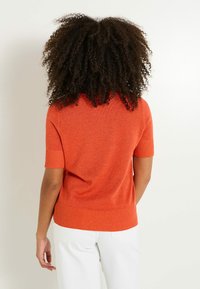 Suéter naranja de manga corta con cuello y dobladillo acanalados, que presenta un acabado de punto texturizado, combinado con pantalones blancos.