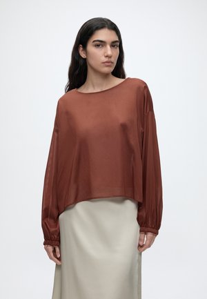 Mujer con cabello largo y oscuro viste una blusa suelta de manga larga color óxido y una falda beige claro, de pie contra un fondo liso.