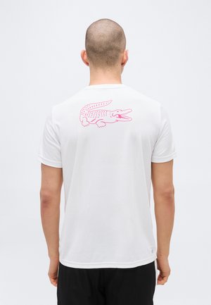 Man with shaved head wearing white t-shirt with large pink crocodile graphic on upper back, standing against plain light background.