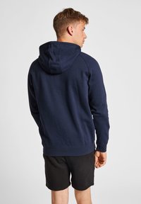 Felpa blu navy con tasca a marsupio e cappuccio regolabile, realizzata in morbido tessuto, con polsini e orlo a costine. Indossata sopra pantaloni corti neri.
