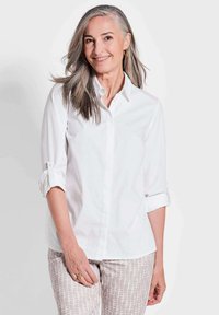 Weißes Hemd mit gekrempelten Ärmeln, aus sanftem Baumwollstoff. Dazu hellgemusterte Hose in neutralen Farbtönen.