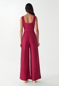 Ženska z dolgimi temnimi lasmi, oblečena v brezrokavnik burgundske barve s širokimi hlačami, stoji pred enobarvno belo ozadje.