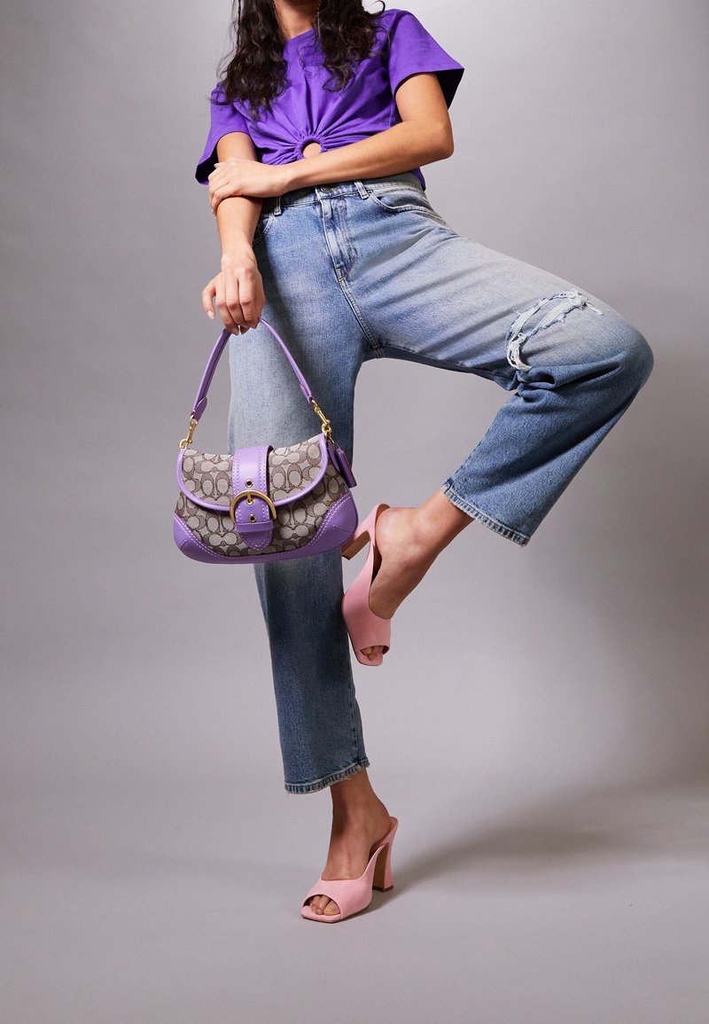 Lilla håndtaske med en mønstret stofkrop, lavendel detaljer og guld hardware. Modellen bærer lyseblå slidte jeans og rosa højhælede mule-sko.