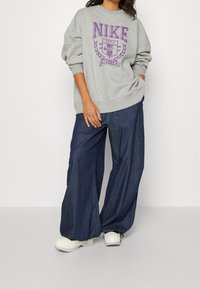 Grå sweatshirt med lila Nike-logotyp och texten 1972, kombinerad med lösa mörkblå denimbyxor och vita sneakers. Mjuk, avslappnad passform.