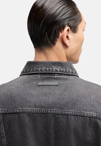 Schwarze Jeansjacke mit kontrastierenden Nähten, ausgestattet mit einem Kragen und einem rechteckigen Logo-Patch auf der Rückseite unterhalb des Nackens.