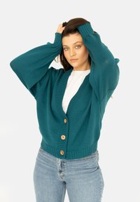 Skrócona kardigan w kolorze turkusowym, dzianinowy, z głębokim dekoltem w kształcie V, oversize'owymi rękawami oraz zapięciem na drewniane guziki. Rybkowe mankiety i dół dodają tekstury. Noszony na białej bluzce.