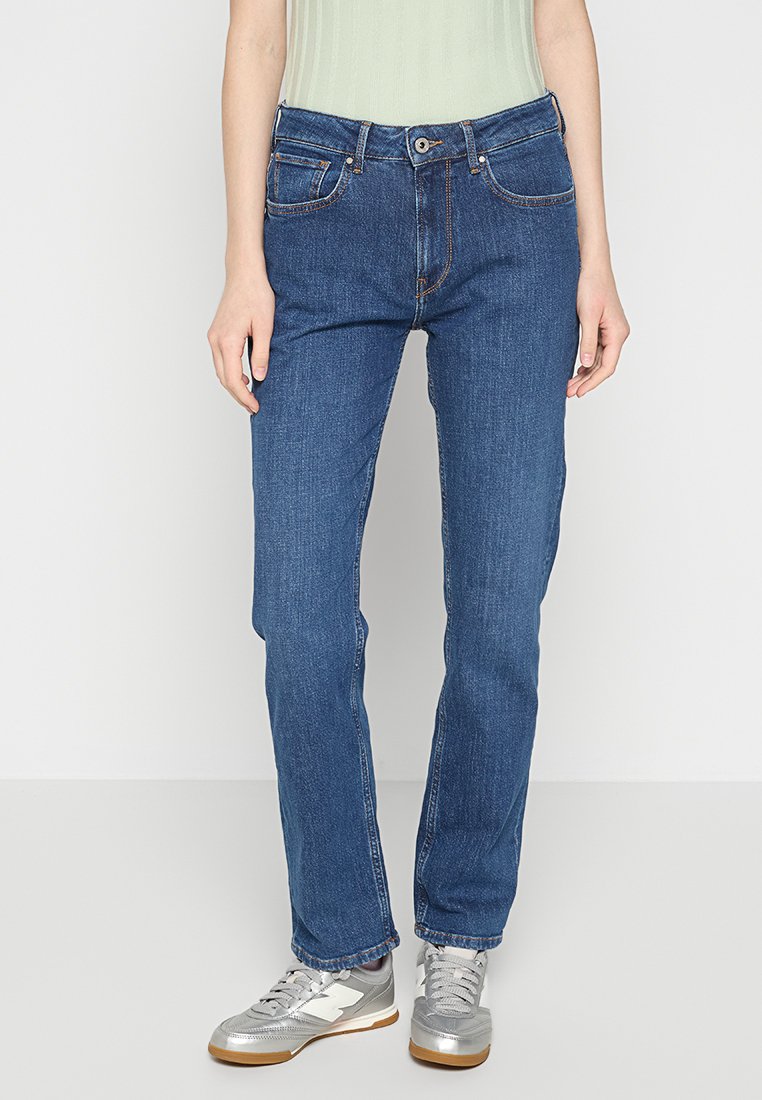 Blaue Denim-Jeans mit geradem Bein, fünf Taschen und orangen Nähten, kombiniert mit silbernen Sneakers und einem hellgrünen Oberteil.
