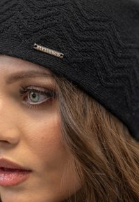 Beanie nero a maglia con un motivo a zigzag testurizzato e una targhetta metallica, progettato per calore e stile, con una vestibilità aderente.