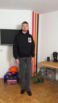 Zwarte hoodie met een voorzak en logo, gecombineerd met grijze spijkerbroek. De achtergrond toont kleurrijke strepen op de muur en een houten vloer.