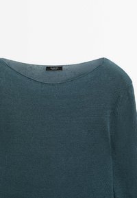 Pull tricoté sarcelle avec encolure bateau et manches longues, montrant le haut de la poitrine et des épaules.