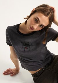 Mørkegrå T-shirt med korte ærmer, sort ribbet halsudskæring og en broderet grafisk design i sort og rød, der viser en figur med stjerner.