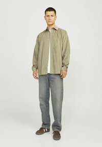 Cămașă lungă cu mâneci, de culoare verde măslin deschis, purtată peste un tricou alb. Asortată cu blugi largi de culoare albastră și adidași maro cu accente albe.