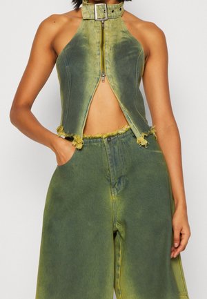 Zielonkawo-żółty zniszczony top na halter z zamkiem z przodu i kołnierzem z klamrą, zestawiony z pasującymi szerokimi spodniami z ręką w kieszeni.