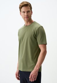 Oliwkowa koszulka z krótkim rękawem z miękkiego materiału, z okrągłym dekoltem i luźnym krojem, w połączeniu z ciemnoniebieskimi spodniami.