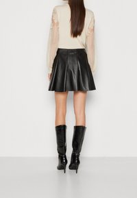 Schwarzer Lederrock mit Falten, kombiniert mit einem cremefarbenen Oberteil mit transparenten, strukturierten Ärmeln. Das Modell trägt kniehohe, glänzende schwarze Stiefel.