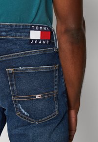 Jeans denim w ciemnoniebieskim kolorze o postarzanym wyglądzie. Zawierają łatkę TOMMY JEANS z czerwono-biało-niebieskim wzorem na tylnej kieszeni.