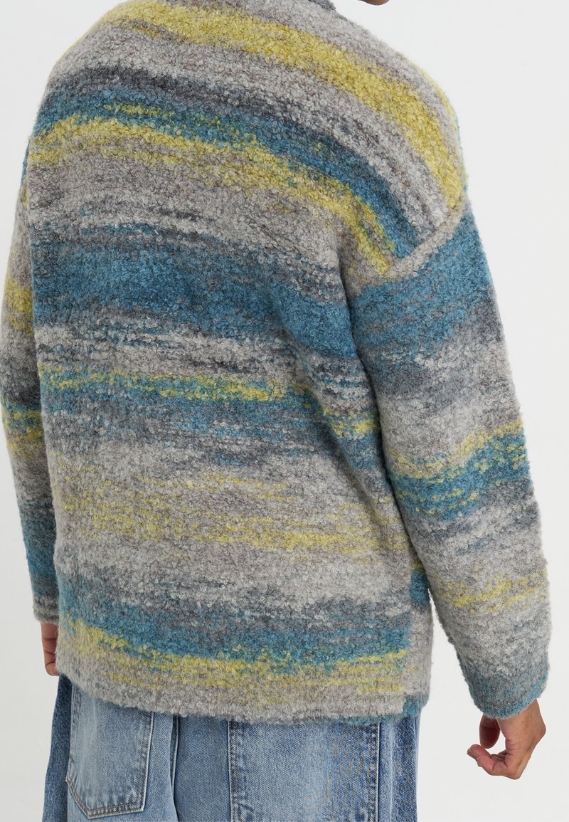 Pull en tricot gris avec des rayures bleues, jaunes et grises ; surface texturée et moelleuse ; coupe ample ; col et poignets côtelés ; manches mi-longues.