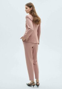 Costume ajusté rose clair avec une veste cintrée et un pantalon slim. Texture douce avec des détails de boutons. Associé à des escarpins noirs à bout pointu.