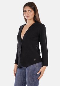 Cardigan nero con scollatura a V profonda, maniche lunghe e design aderente. Presenta un piccolo dettaglio con logo all'orlo. Indossato sopra una maglietta chiara.