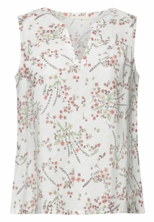 Femme portant un top blanc sans manches à motifs floraux, un pantalon large orange vif et des sandales blanches, debout devant un fond orange.