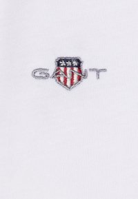 Hvit stoff med et lite brodert skjoldlogo med røde og blå striper, stjerner og teksten "GANT" i grått.