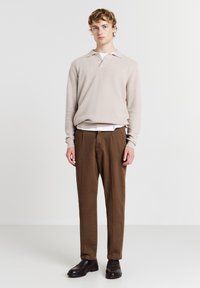 Junger Mann steht, trägt einen beigen Strickpullover, ein weißes Unterhemd, braune Hosen und dunkelbraune Slipper vor einem schlichten weißen Hintergrund.