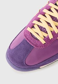 Tênis roxo com uma combinação de material em camurça e nylon, detalhes texturizados, sola de borracha e atacadores amarelos para contraste.