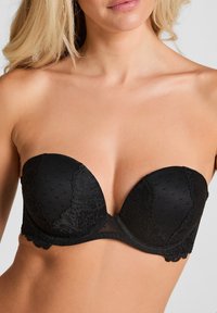 Vrouw in een zwart strapless kant-bh, zichtbaar is de bovenste torso en decolleté met gedeeltelijk zichtbaar blond haar.