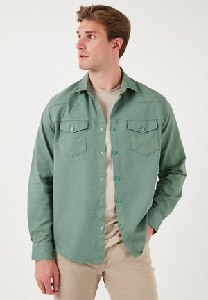 Green button-up shirt made of cotton, featuring two chest pockets, snap buttons, and a relaxed fit. Background is plain white.