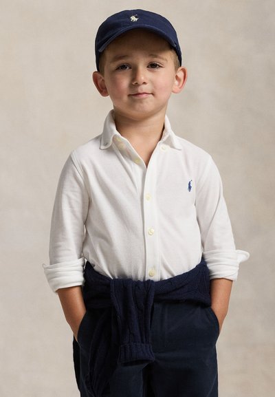 Niño joven que lleva una camisa blanca abotonada, gorra azul marino y suéter azul marino atado alrededor de la cintura con las manos en los bolsillos, de pie frente a un fondo sencillo.