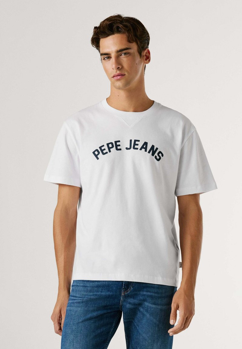 Jeune homme portant un t-shirt blanc Pepe Jeans et un jean bleu, debout devant un fond clair uni.